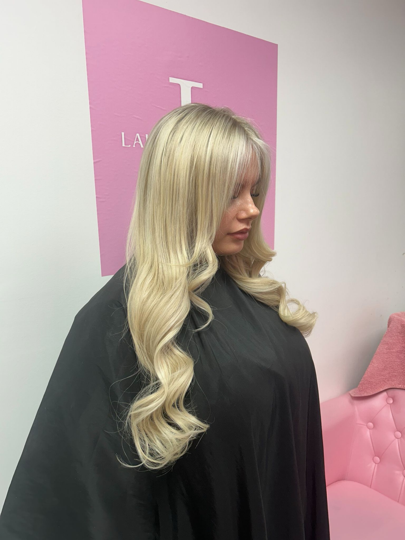 A person with long wavy blonde hair, wearing a black gown, standing in a room with a pink chair and a sign in the background.