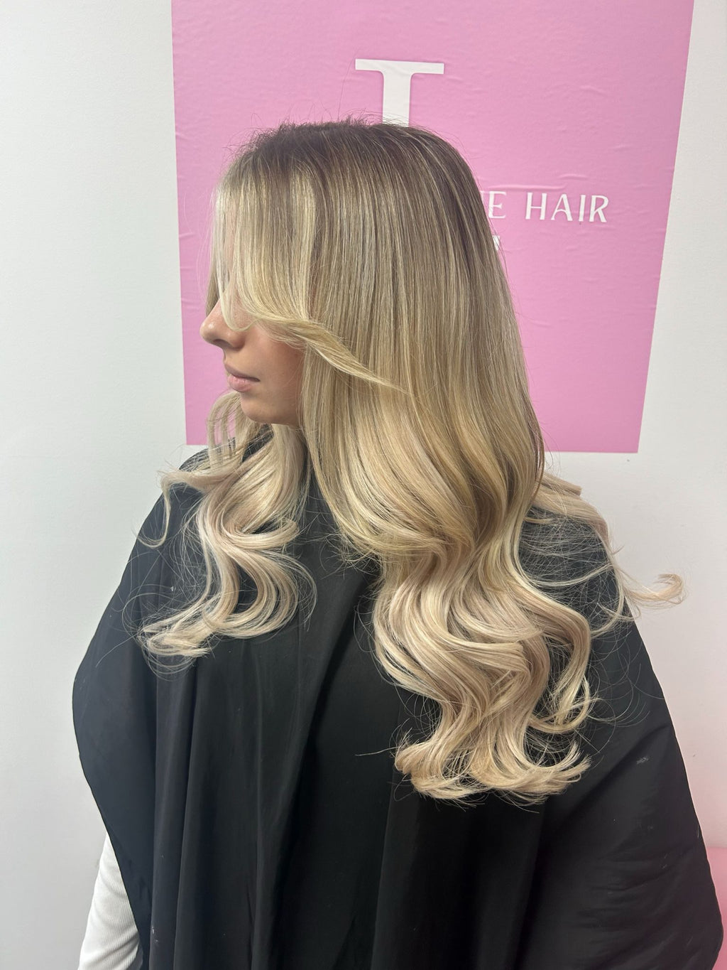 A person with long wavy blonde hair, wearing a black garment, standing in front of a pink wall.